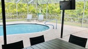 Un espace piscine offrant une vue dégagée sur la piscine, un téléviseur fixé sur un poteau, ainsi qu’une table et des chaises disposées pour les repas.