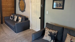 A cozy living room with two grey sofas, each with patterned cushions, a wooden ceiling, a white door, and a framed picture on the wall. - Cabin Retreat in Forest Lakes ... 2 hours from the Valley (Forest Lakes Estates)