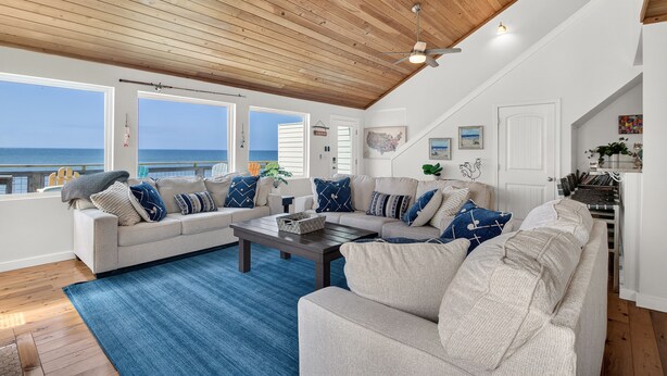 A spacious living room with a wooden ceiling, large windows overlooking the ocean, a beige sofa set, and a coffee table.