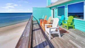 A wooden deck with colorful Adirondack chairs facing a turquoise building with large windows overlooking a beach.