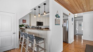 A kitchen with white cabinets, a microwave, and a stainless steel refrigerator. There are bar stools and a hanging light fixture.