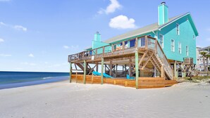 A turquoise beachfront house with a wooden deck and stairs leading to the main floor.