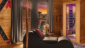 A modern log cabin interior with a black sofa, a view of a sunset through a door, and a wooden staircase.