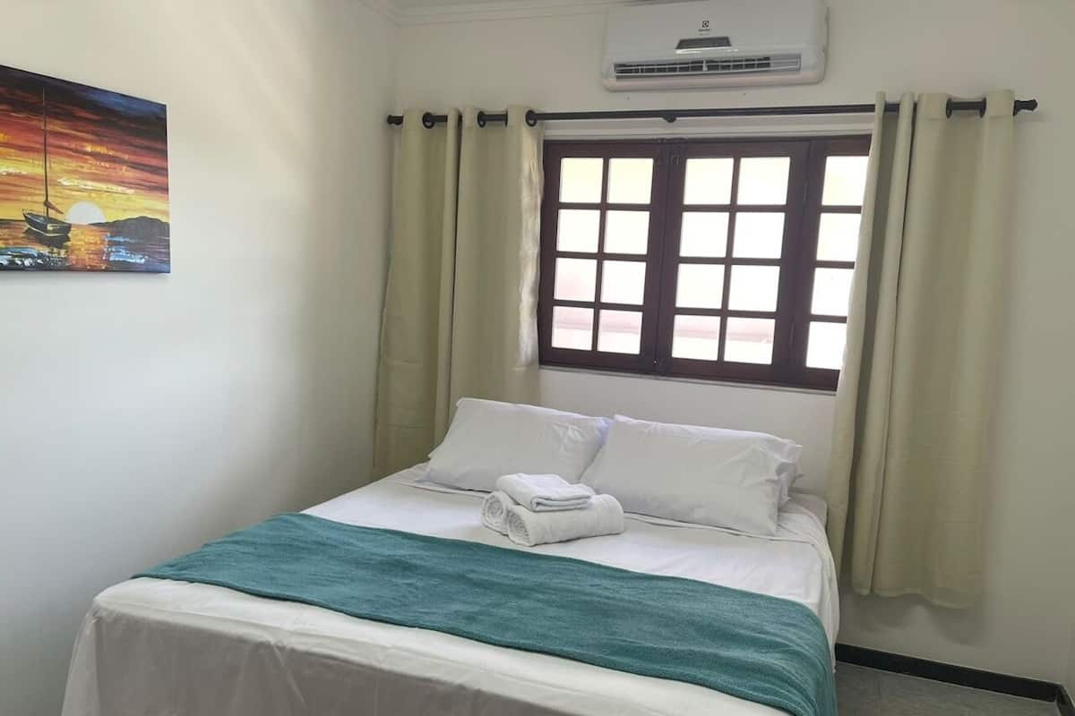 Una cama bien hecha con sábanas blancas y una manta color turquesa, una ventana con cortinas y un equipo de aire acondicionado arriba.