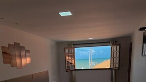 A hotel room with three beds arranged in a row, a large window with a view of the beach, and a modern light fixture on the wall. - Pousada La Riviera (Maragogi)
