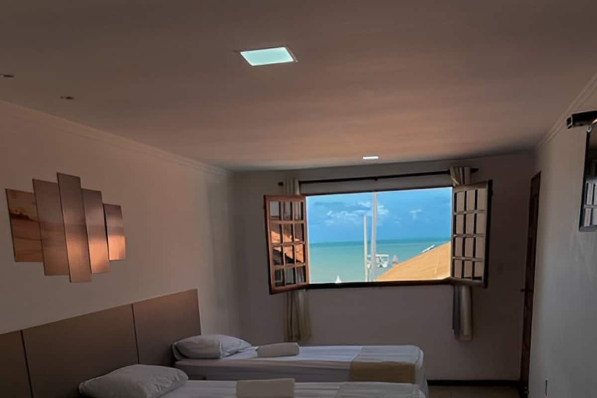 Habitación de hotel con tres camas alineadas, un ventanal con vistas a la playa y una lámpara moderna en la pared.
