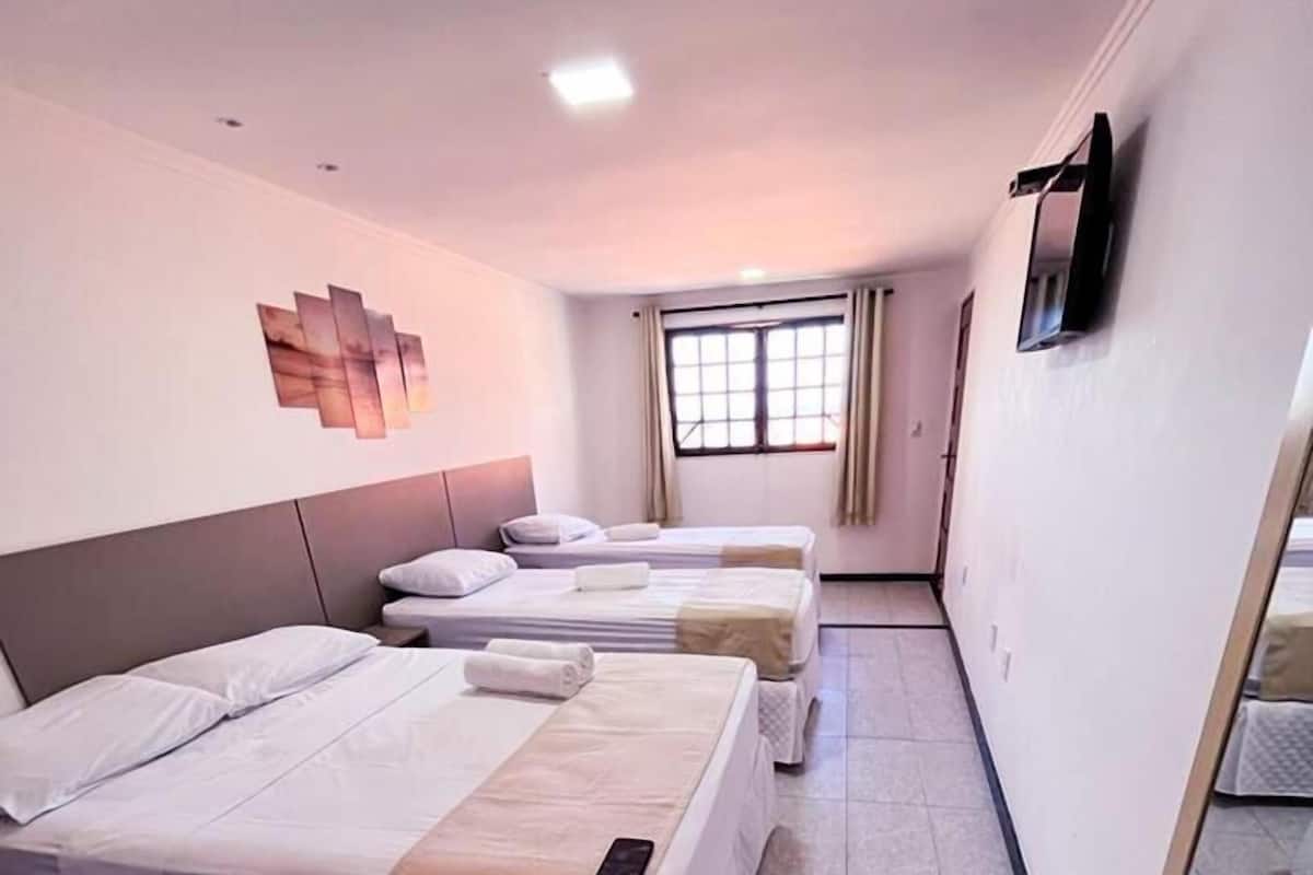 Habitación de hotel con tres camas, un televisor montado en la pared y una ventana con cortinas.