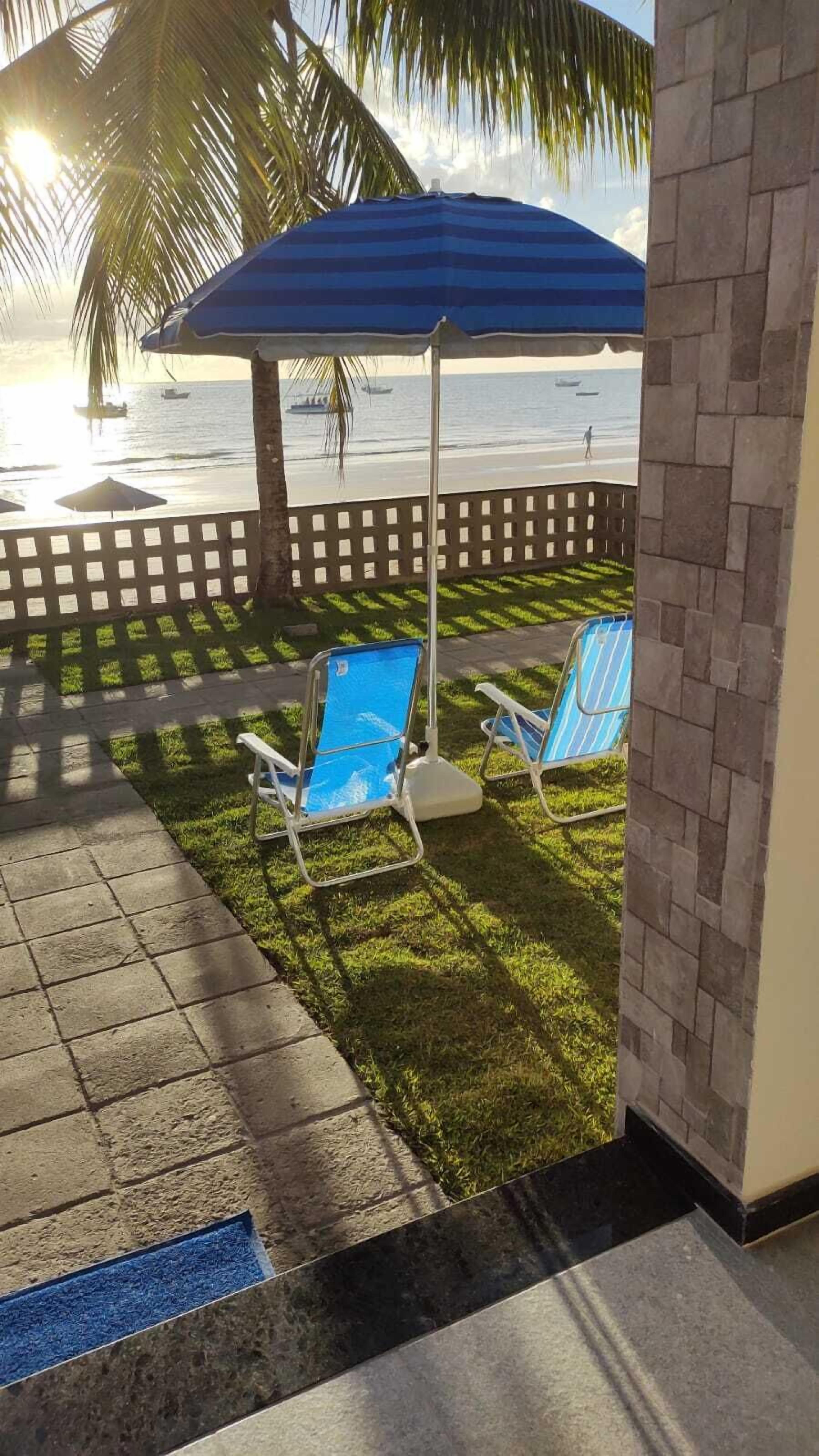 Un área junto a la piscina con dos sillas reclinables y una sombrilla azul, una palmera y vista al océano.