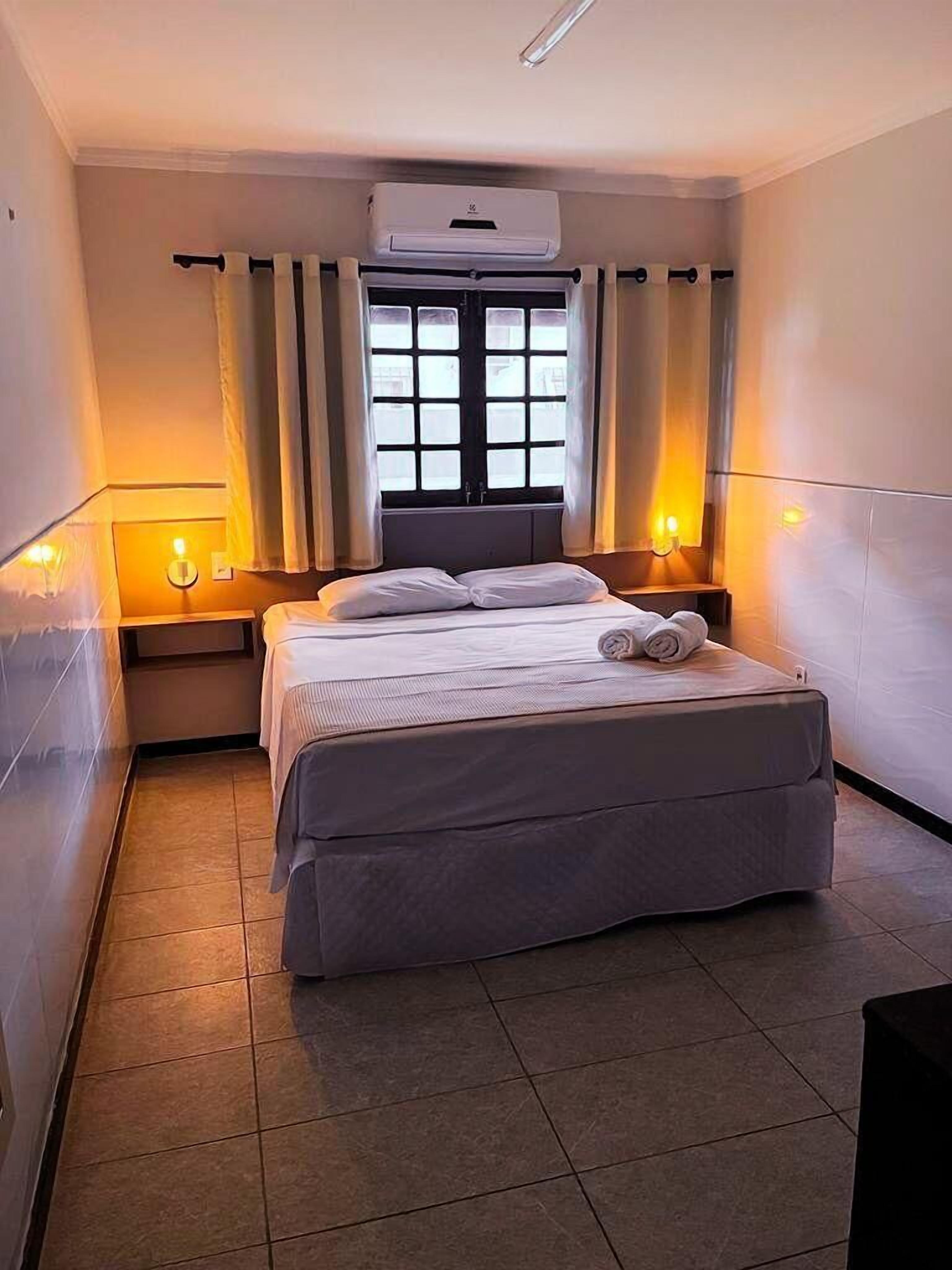 Habitación de hotel con cama, dos mesitas de noche, ventana con cortinas, unidad de aire acondicionado y lámpara fijada a la pared.