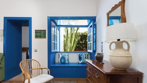 Une pièce avec un banc intégré près d’une fenêtre bleue, une chaise en osier, une commode en bois et un grand vase.