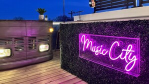 A neon sign reading "Music City" against a backdrop of a wooden deck and a wall with foliage. - Rooftop Bar ★ Downtown ★ Brand New (Nashville)