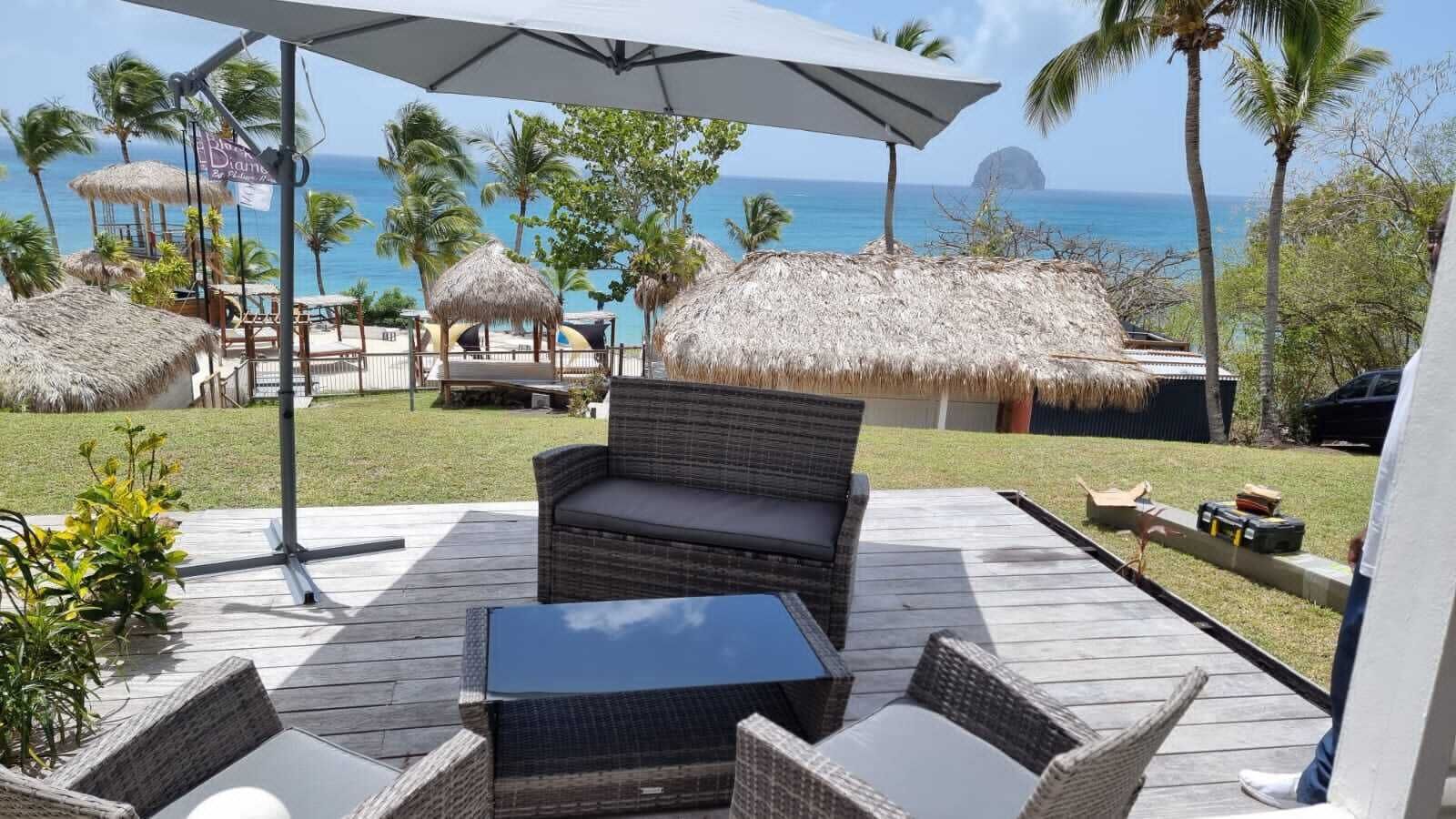 A patio with wicker furniture, a table, and a large umbrella. There are thatched umbrellas and palm trees in the background.