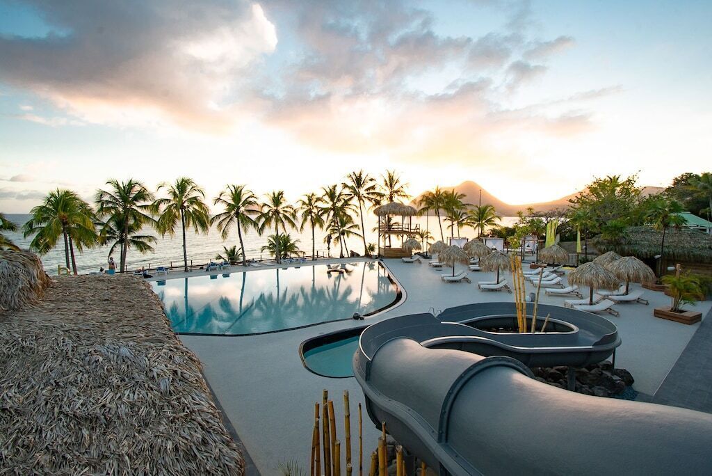 Un espace piscine dans un complexe hôtelier, agrémenté de palmiers, de chaises longues et offrant une vue sur le coucher de soleil.