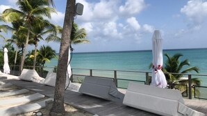 A beachside lounge area with white lounge chairs and umbrellas, palm trees, and a view of the ocean. - 3-person studio deck 15m2 facing Le Diamant (Le Diamant)