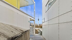 Ein schmaler Außenkorridor mit Holzdeck, Glaswand und Blick auf eine Stadtlandschaft mit einem Turm.