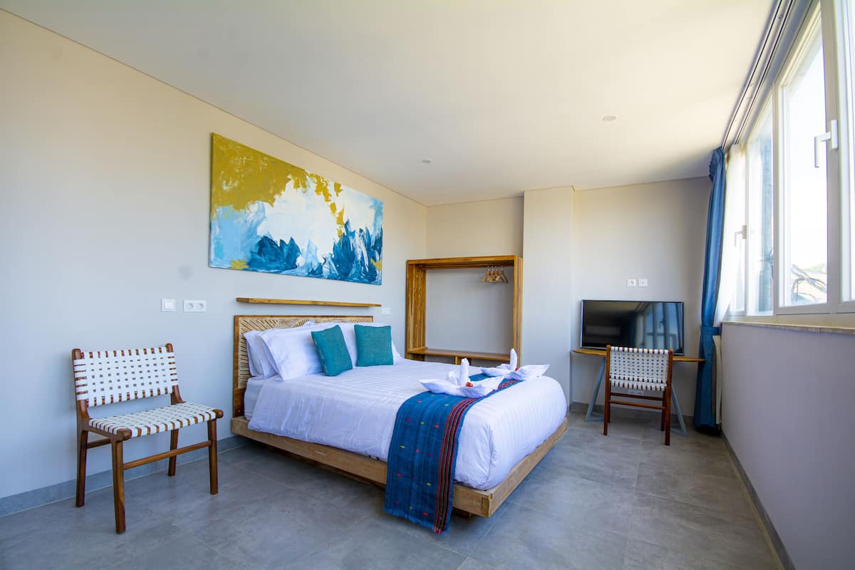 A modern hotel room with a large bed, two wooden chairs, a TV, and a painting on the wall.