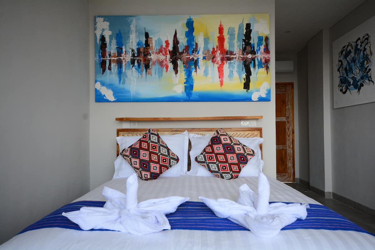 A hotel room with a large bed, two patterned pillows, a blue-striped bedspread, and a painting above the bed.