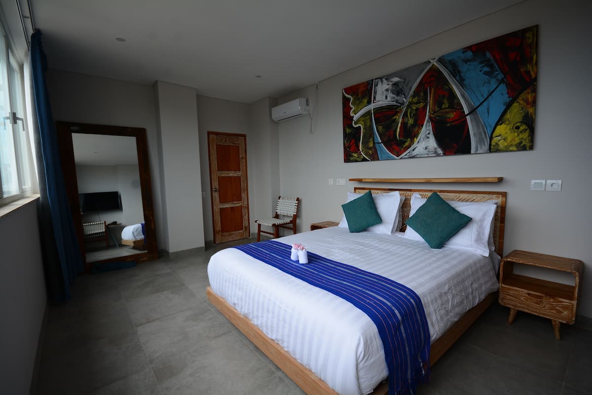 A neatly arranged bedroom with a large bed, a wooden nightstand, a chair, and a colorful abstract painting on the wall.