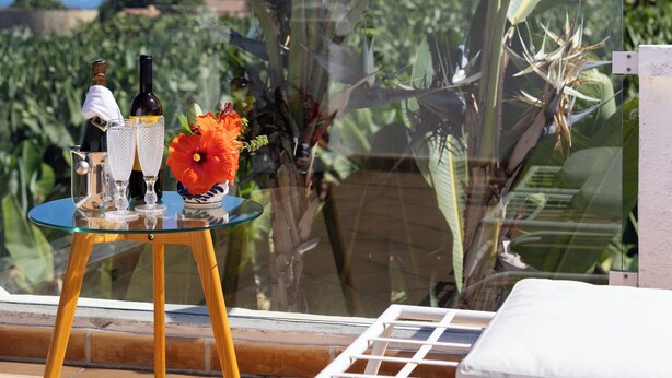 Ein Glastisch mit Flasche und Gläsern, eine Vase mit einer Blume sowie ein Stuhl auf einer Terrasse mit Blick auf Bananenstauden und Hügel.