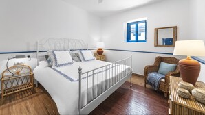 Ein ordentlich eingerichtetes Schlafzimmer mit einem Metallbettgestell, weißer Bettwäsche, einem Korbstuhl, einem Nachttisch aus Holz und einem Fenster mit blauen Fensterläden.