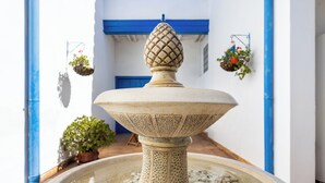Ein Steinfountain mit dekorativer Spitze, umgeben von einer runden Schale gefüllt mit Kieselsteinen, vor einer weißen Wand mit blauen Akzenten und hängenden Blumenkörben.