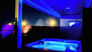 Una habitación con un jacuzzi grande e iluminado, un televisor montado en la pared y una estructura de techo de madera.