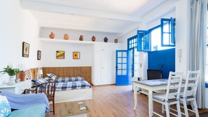 Ein moderner, hell erleuchteter Raum mit einem Bett, einem Schreibtisch und einer Essecke. Regale mit Dekorationselementen und ein großes Fenster mit blauen Fensterläden.