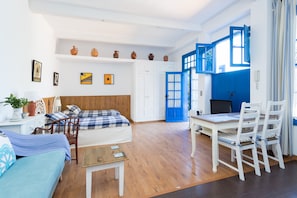 Una camera moderna e luminosa con un letto, una scrivania e un'area pranzo. Sono presenti mensole con oggetti decorativi e un'ampia finestra con persiane blu.