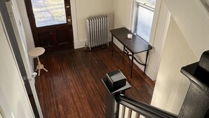 A staircase with a wooden handrail and white balusters, leading to a room with a dark wooden floor, a radiator, and a small table with a chair.