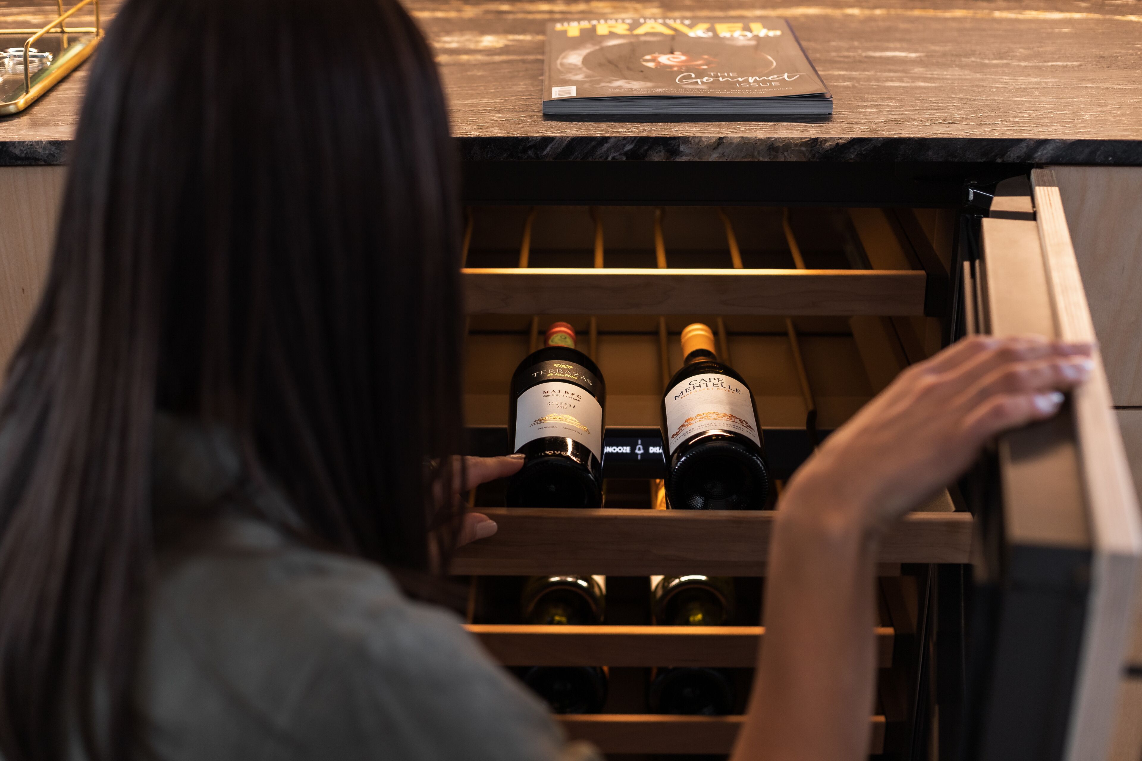 A person opening a wooden wine rack in a room with a book titled "TRAVEL" on the table.