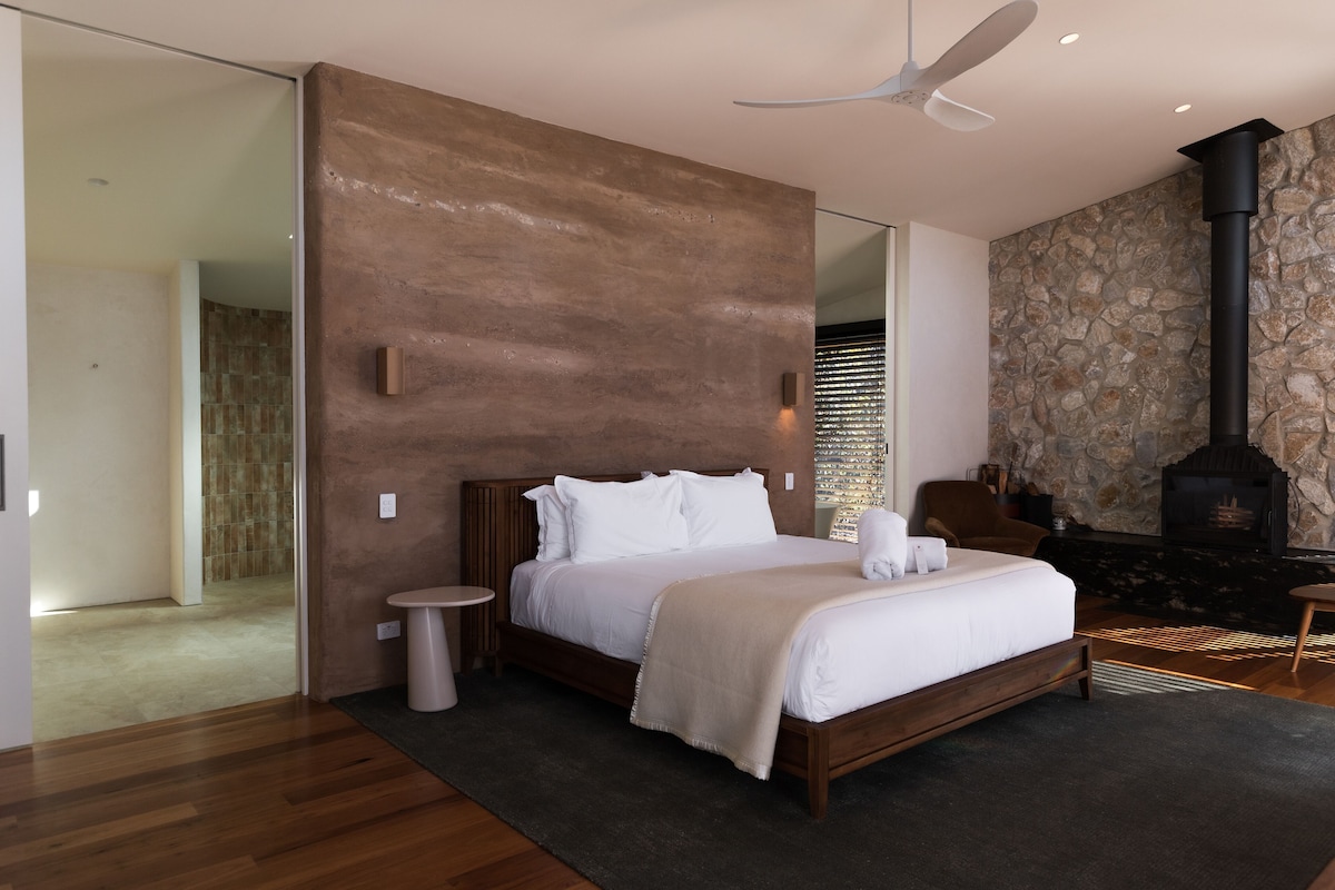 A modern bedroom with a large bed, a stone wall, a fireplace, and a ceiling fan.