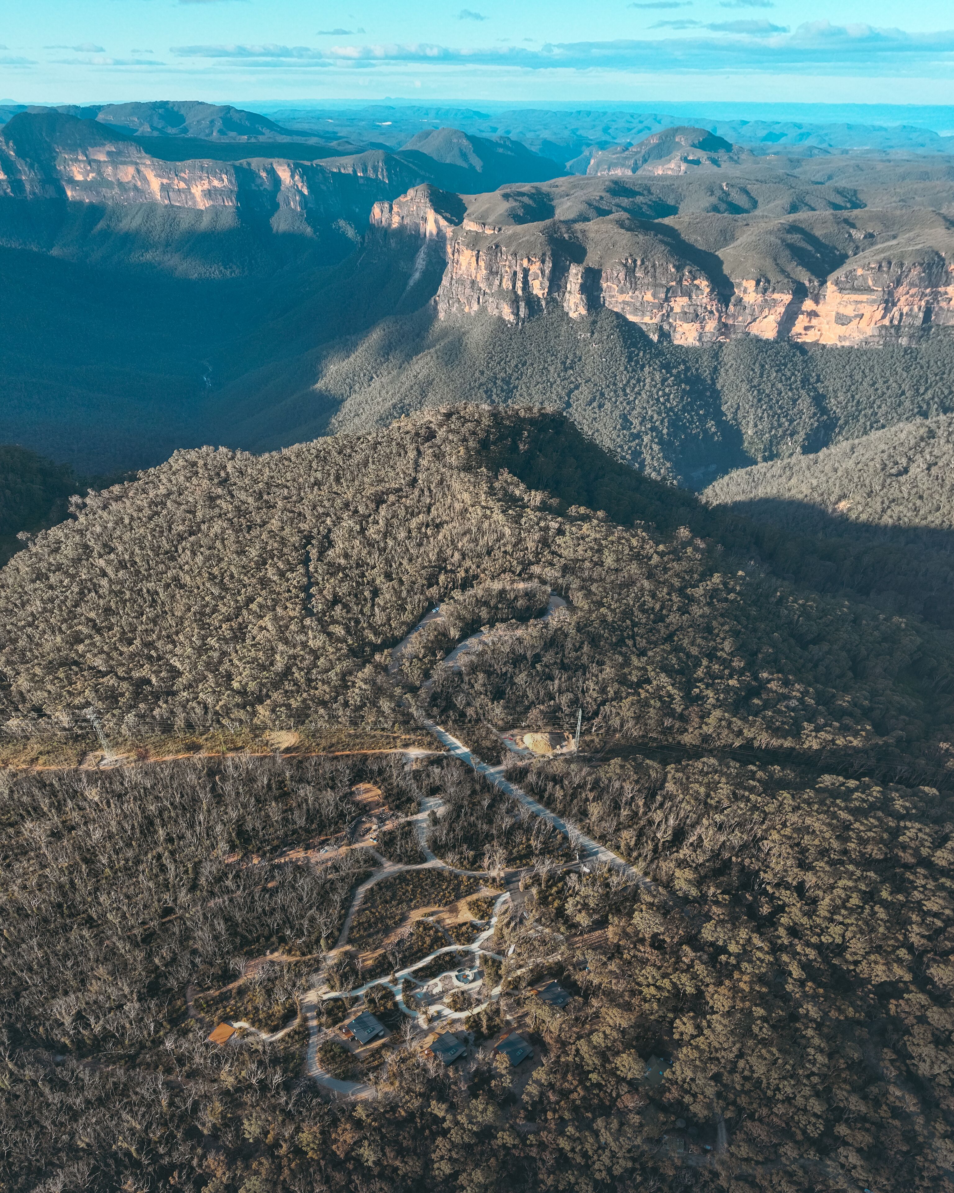 An aerial view of a mountainous area with a winding road, dense forest, and a few buildings.