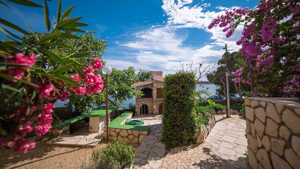 A stone pathway leading to a stone building with a green roof, surrounded by lush greenery and pink flowers.