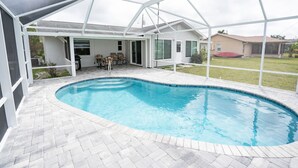 Une piscine couverte avec un espace patio, une maison blanche en arrière-plan et une pelouse verdoyante.