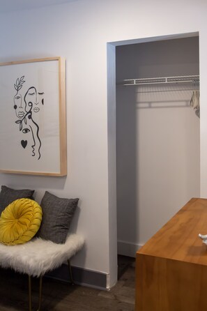 A room with a wooden bench, a framed abstract artwork, a closet with hanging clothes, and a wooden table. - Charming Tiny House W/ Free Parking (Birmingham)
