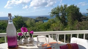 Mesa para dos con una botella, platos, cubiertos y vistas al mar y follaje.