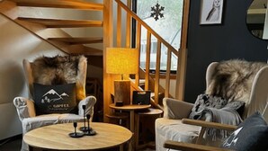 Ein gemütliches Zimmer mit kariertem Boden, einem runden Holztisch, zwei Stühlen mit Felldecken, einer Lampe und einer Treppe mit Holzbalken.