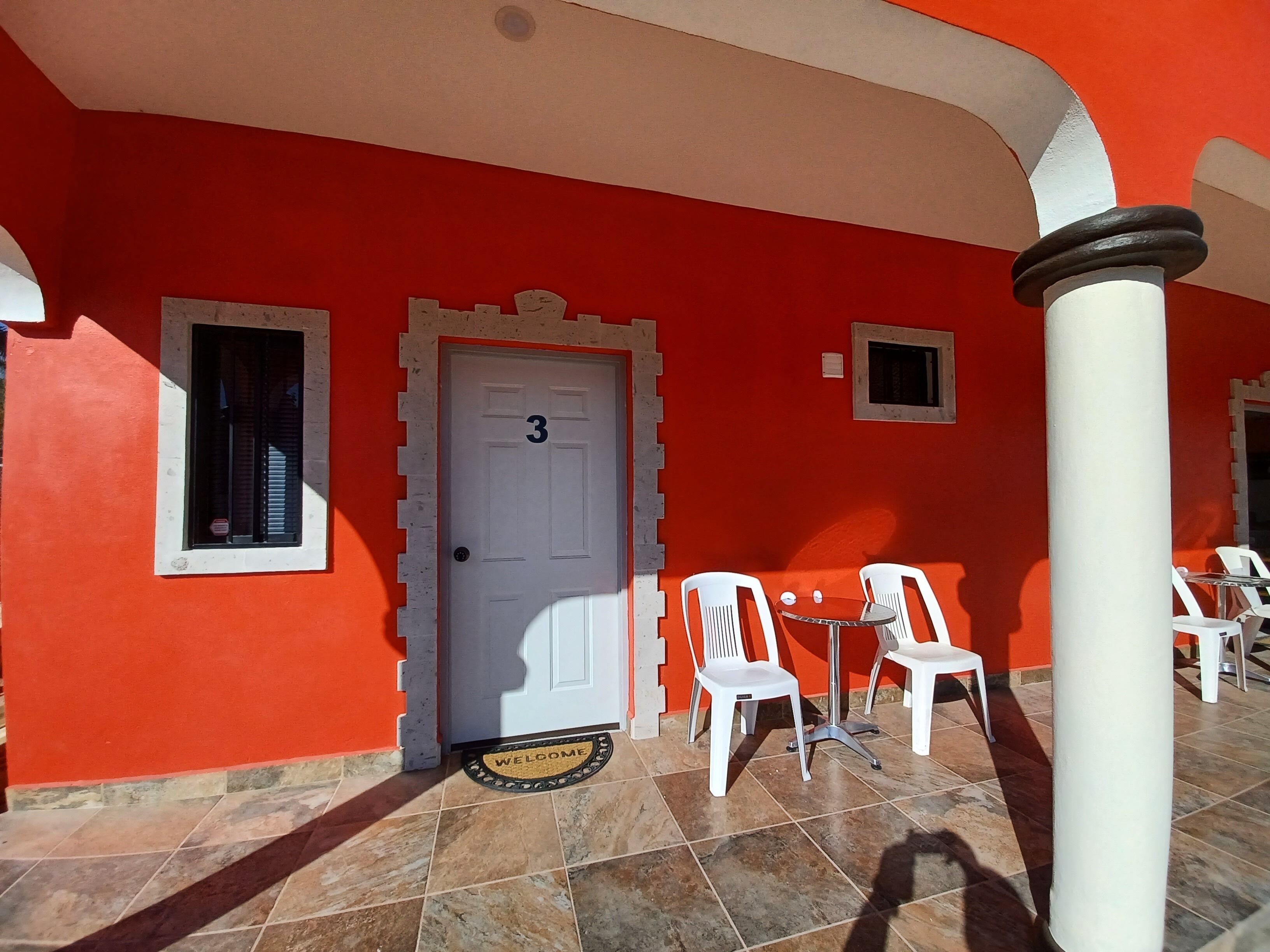 A red building with a white door marked with the number 3, white plastic chairs, and a welcome mat outside.