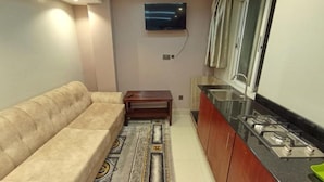 A compact living space with a sofa, a small table, a TV mounted on the wall, a kitchen area with a stove, and a window with curtains. - Decale Palace Hotel (Nairobi)