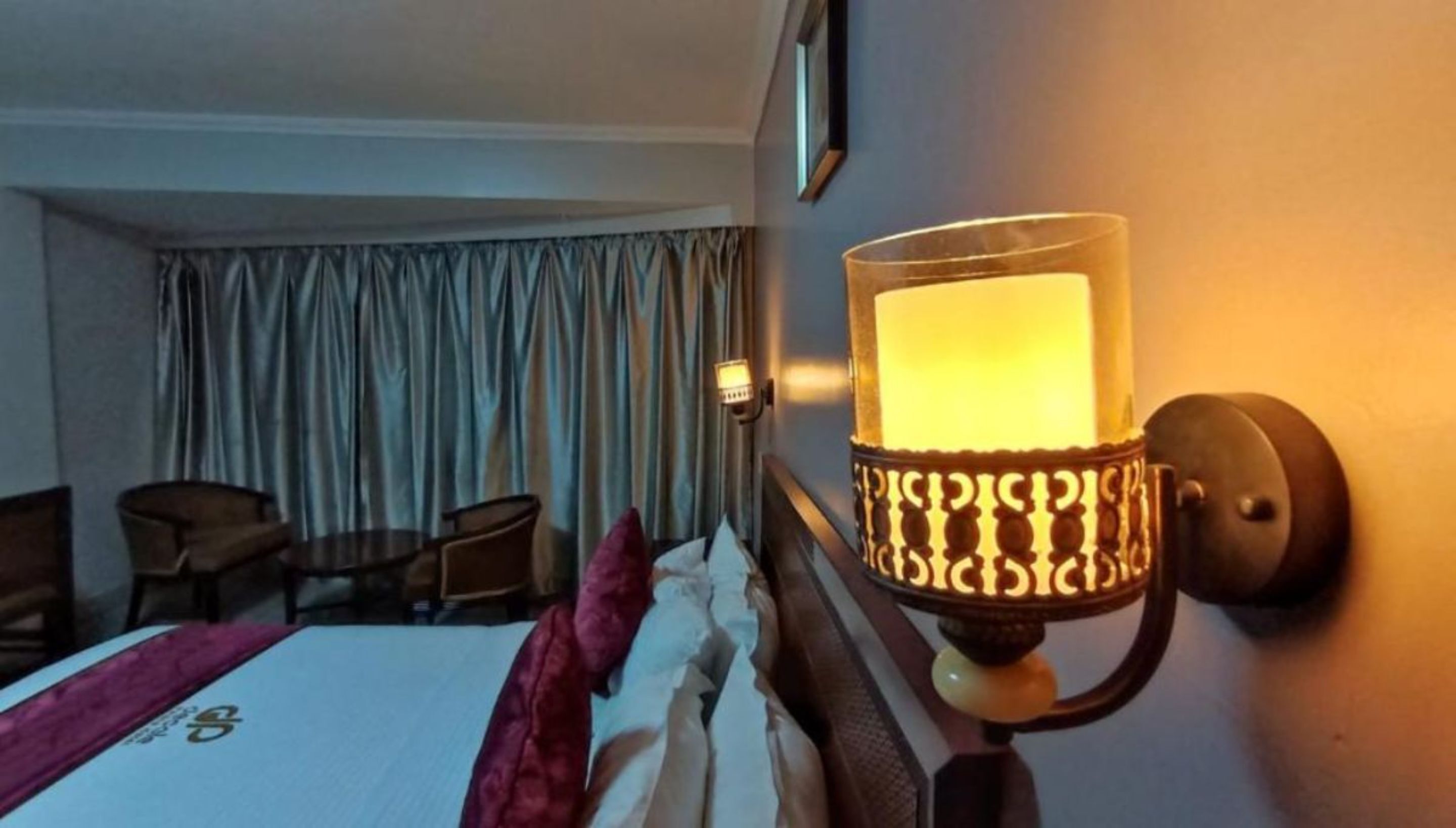 A hotel room with a bed, a nightstand, a lamp, and a view of the outside curtains.