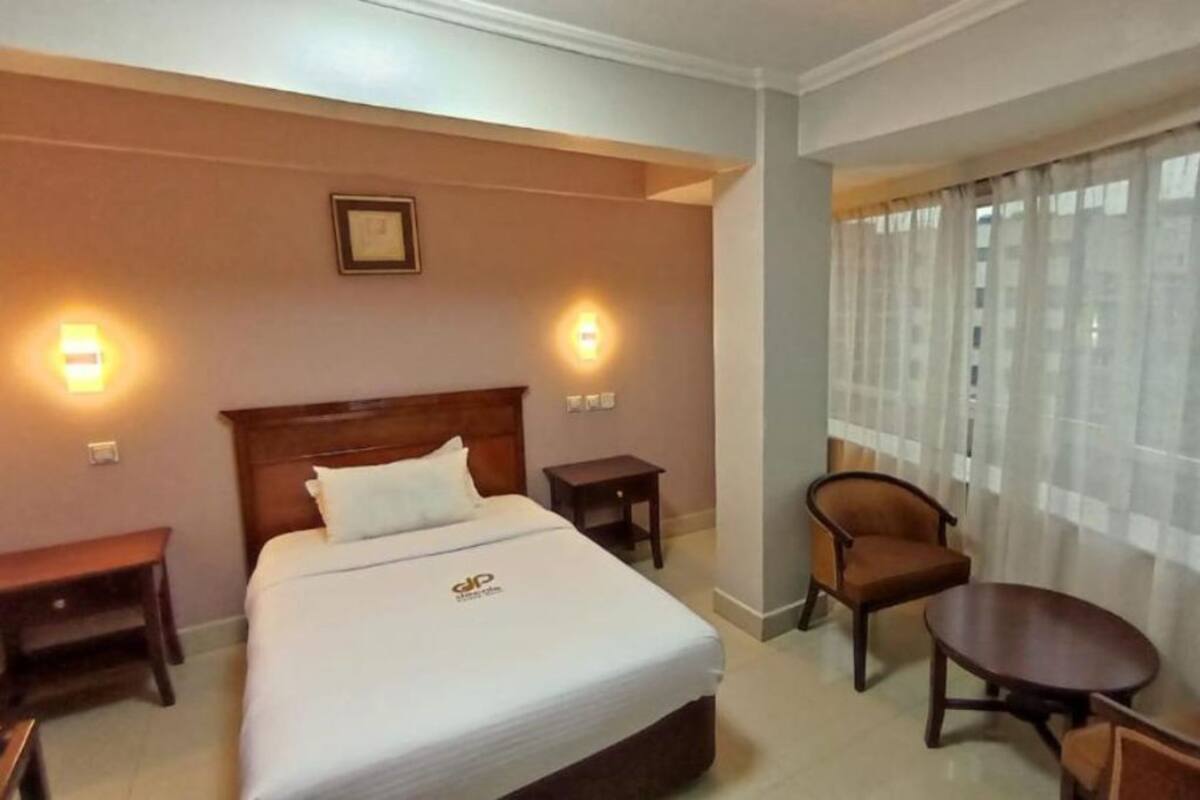 A hotel room with a bed, bedside tables, a chair, a small table, and a window with curtains.