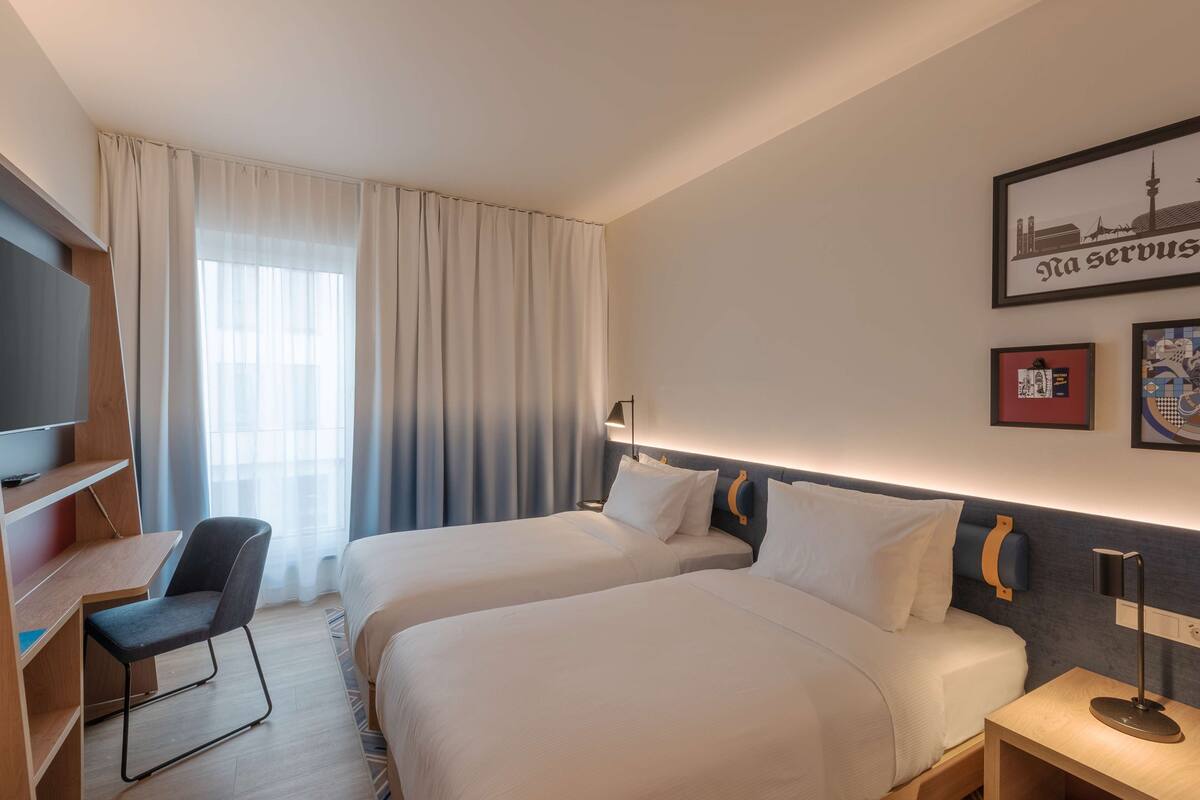 A hotel room with two beds, a desk, a chair, a TV, and artwork on the wall.