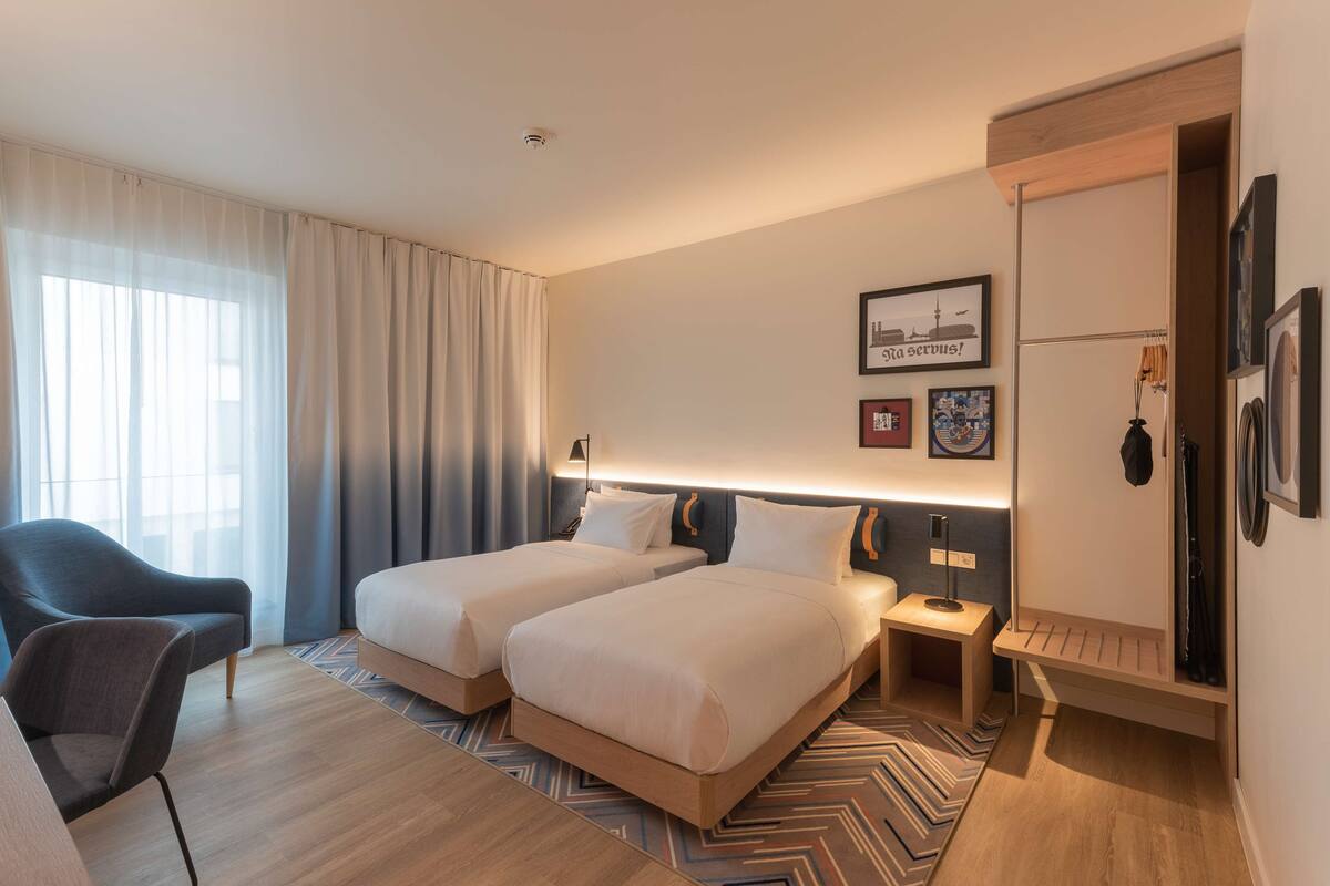 A hotel room with two beds, a chair, a bedside table, a lamp, and a wall with framed pictures.