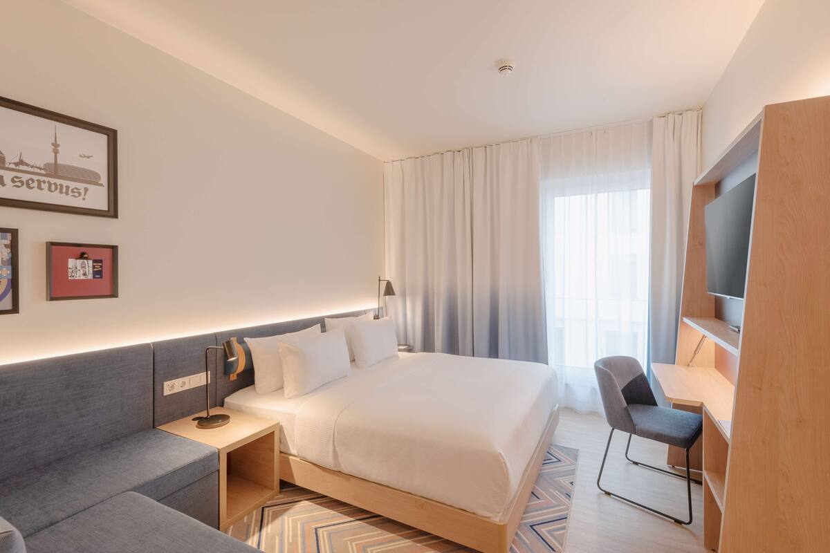A modern hotel room with a bed, a desk, a chair, a TV, and a sofa.
