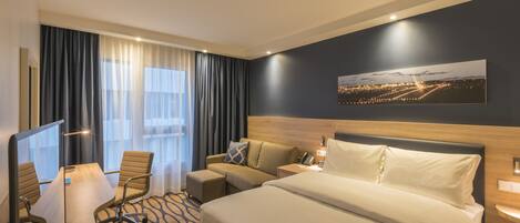 A hotel room with a large bed, a desk with a chair, a small sofa, and a television.