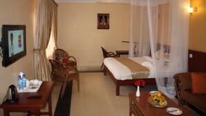 A hotel room with a bed, a desk, a chair, a bedside table, a TV, and a mosquito net. - Kusyombunguo Hotel Lukenya (Athi River)