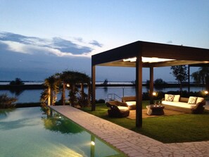 An outdoor pool area with a covered seating area, a view of the water, and a well-maintained lawn. - Beautiful Villa With Private Pool by Beahost (Rosolina)