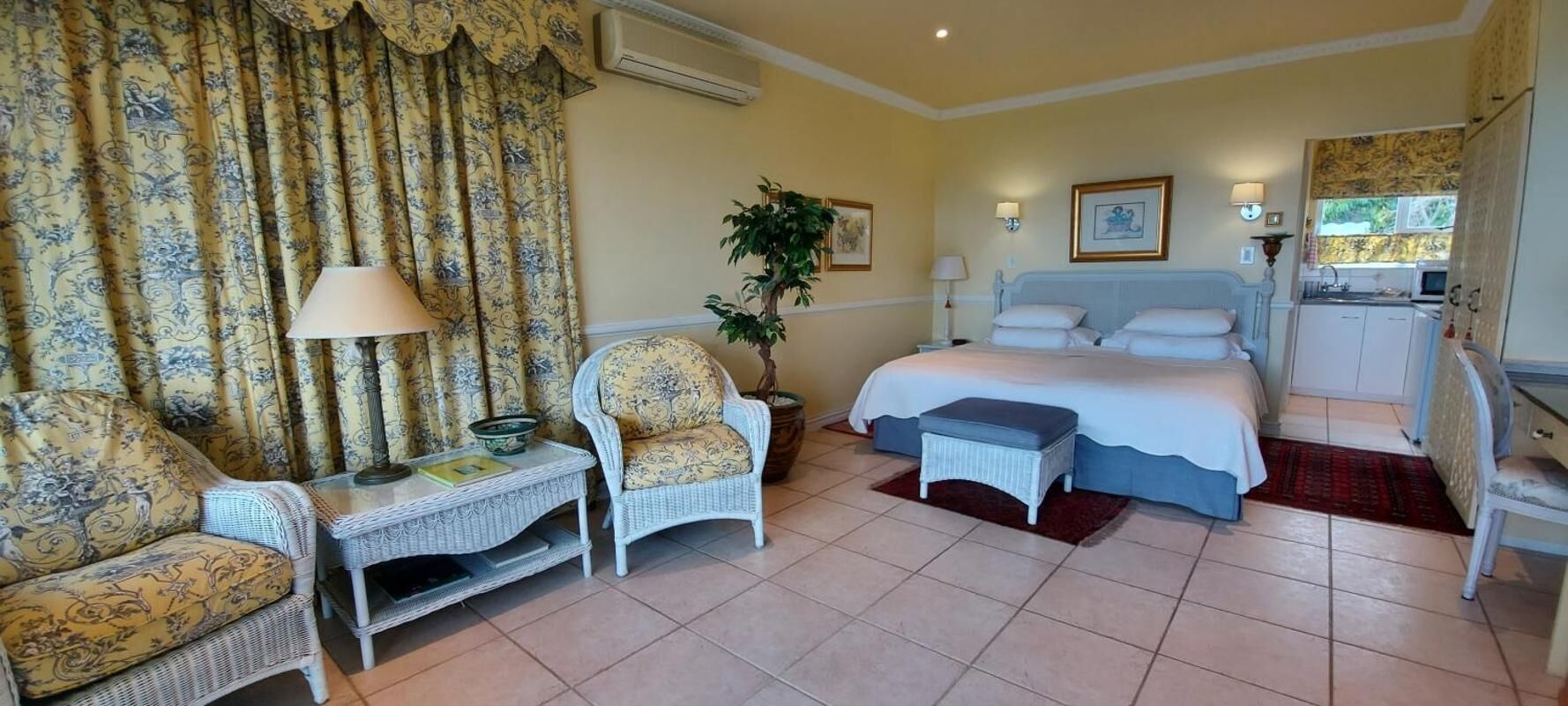 A hotel room with a bed, a chair, a small table, a plant, and a kitchenette.