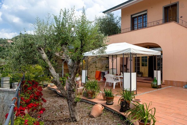 A covered outdoor seating area with a table and chairs, surrounded by potted plants and a tree.