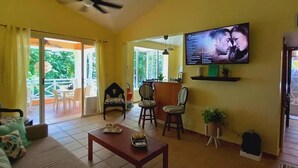 A living room with a sofa, a coffee table, and a TV mounted on the wall. There is a sliding glass door leading to an outdoor area with a patio table and chairs. - "las Brisas, Juan Dolio, 3 Bedrooms, 3 Pools, Jacuzzi, Beach, Golf,polo" (Guayacanes)